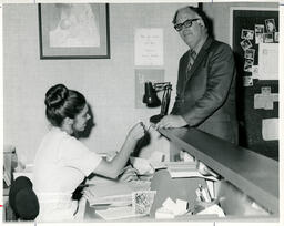Medical office assistant behind a desk assists a customer, ca. 1973