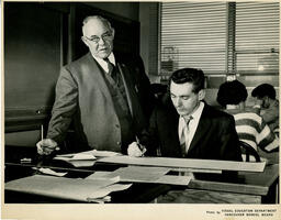 Student writing on a long paper while instructor stands by, ca. 1960