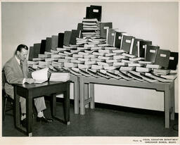 Person at a desk beside a pyramid of books, ca. 1952
