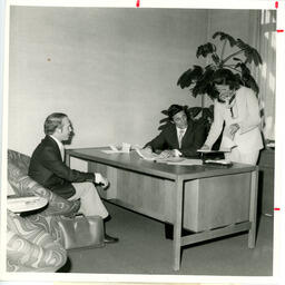 People at desk in an office with one person on a chair
