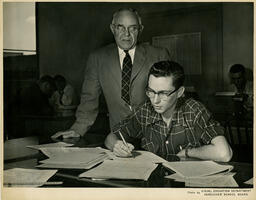 Student writing as instructor watches, ca. 1959