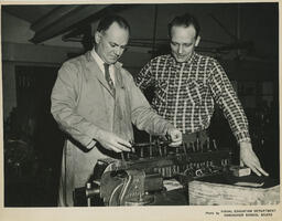 Two people looking at a machine with coils, ca. 1960