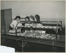 Four students looking at an instructor cutting baked goods, ca. 1949