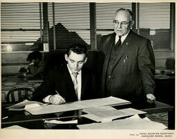 Student writing on paper while instructor watches, ca. 1960