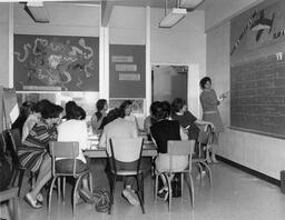 Students at table in classroom looking at teacher, ca. 1970